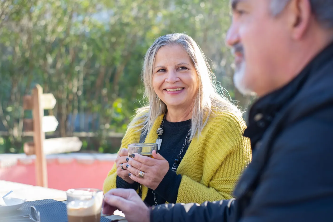 An older woman in a yellow cardigan smiling over a warm drink at a garden table