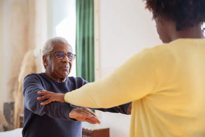 A helper steadying an older man by the arm during a light exercise at home