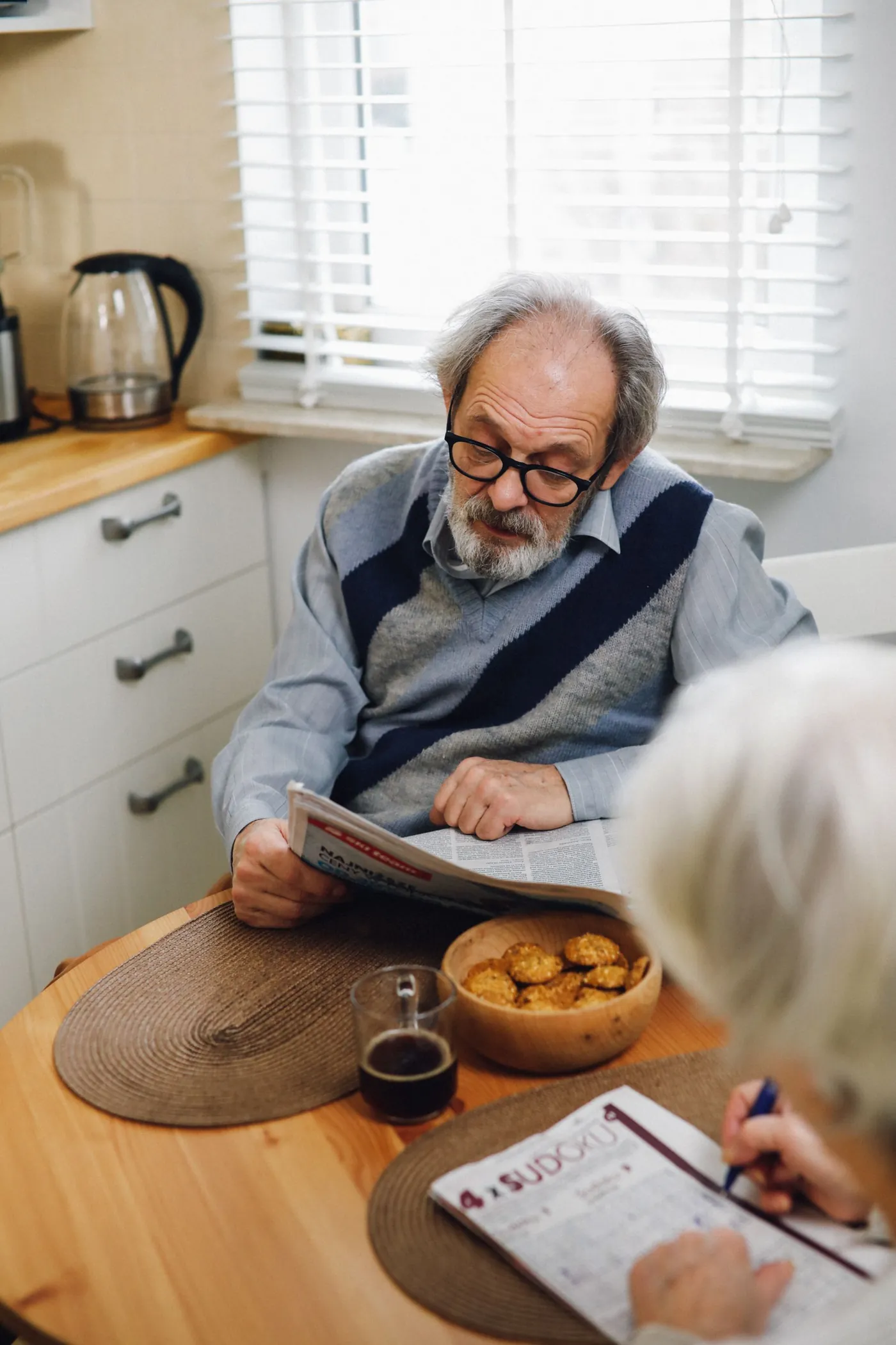 An older man with a newspaper and a cup of coffee at the kitchen table, a sudoku book across from him