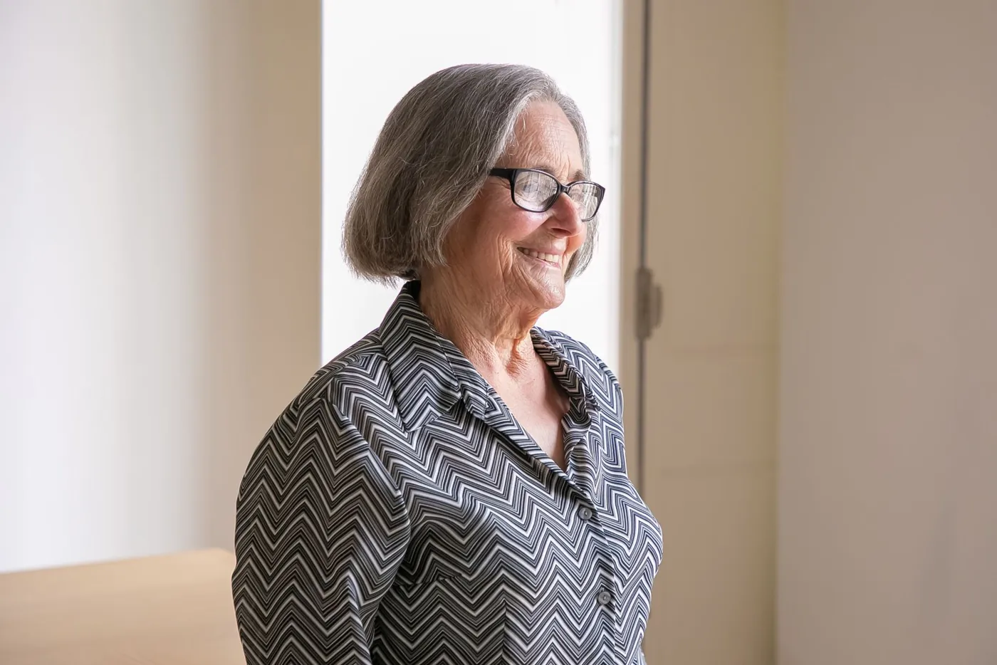 A smiling older woman in glasses looking out of a sunny window at home