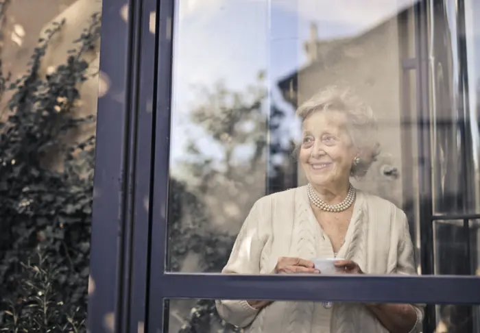 An older woman holding a cup of tea and smiling by her window